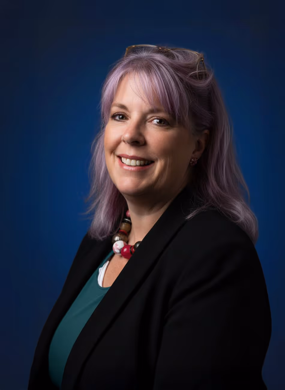 Maven Radd lawyer, Beccy O'Donoghue -  with lavender hair smiles, wearing a black blazer and colorful necklace, against a dark blue background.