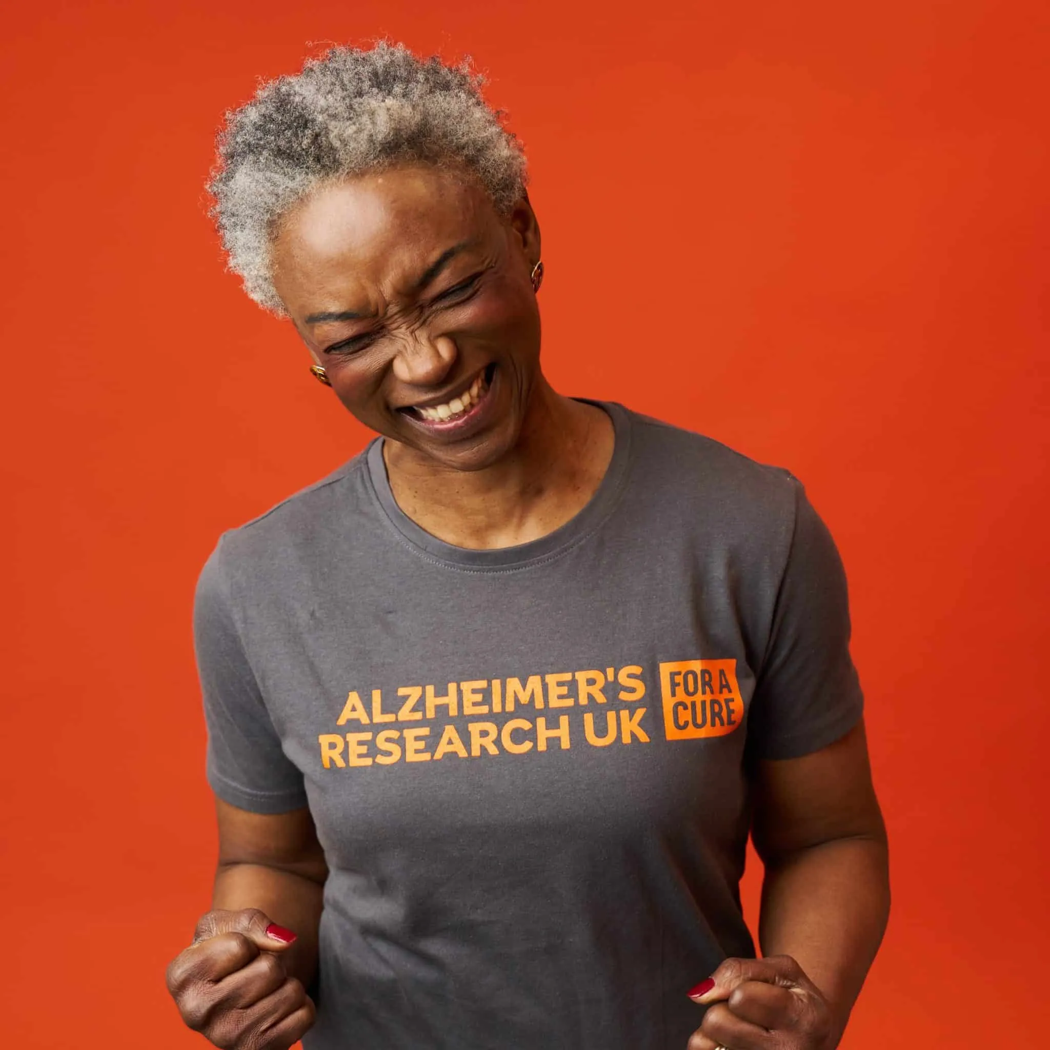 Smiling individual in a gray t-shirt with "Alzheimer's Research UK" printed in orange, posing against a vibrant orange background.
