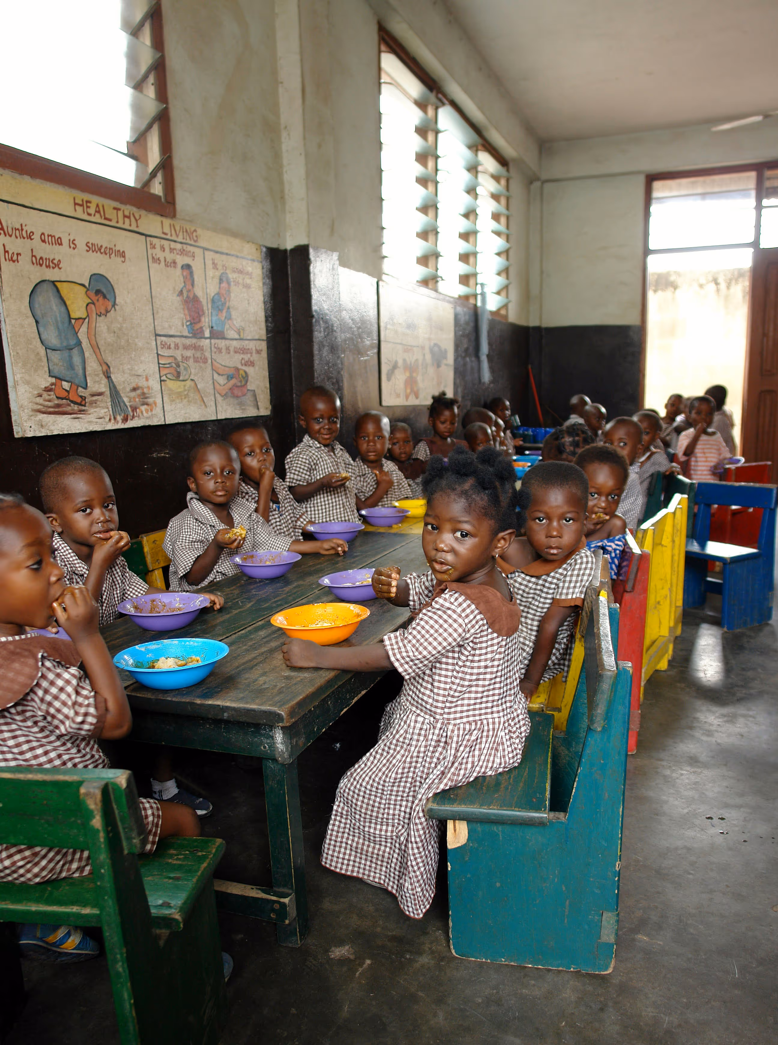 Children in brown checkered uniforms sitting at a table, enjoying meals from colorful bowls in a classroom. Educational posters on the walls.