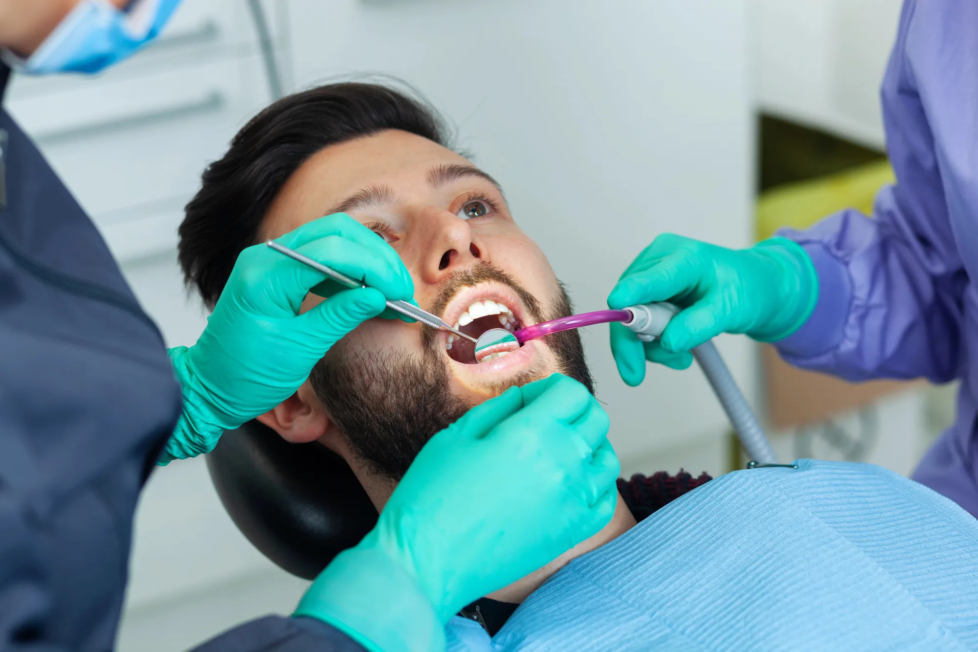 Dentist wearing green gloves examining a man's open mouth using a dental mirror and suction tool.