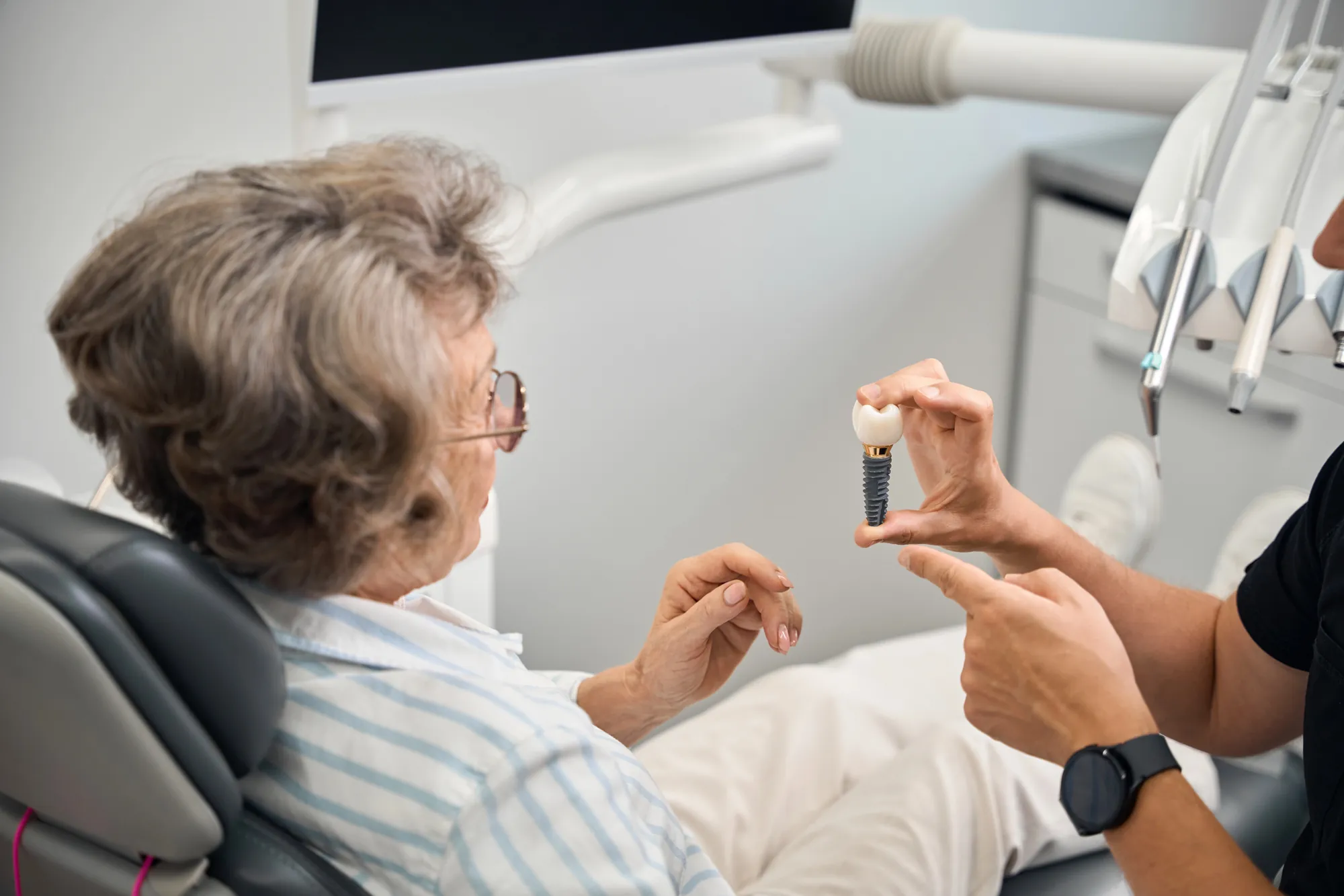 Dentist showing an elderly woman a dental implant model in a dental clinic.