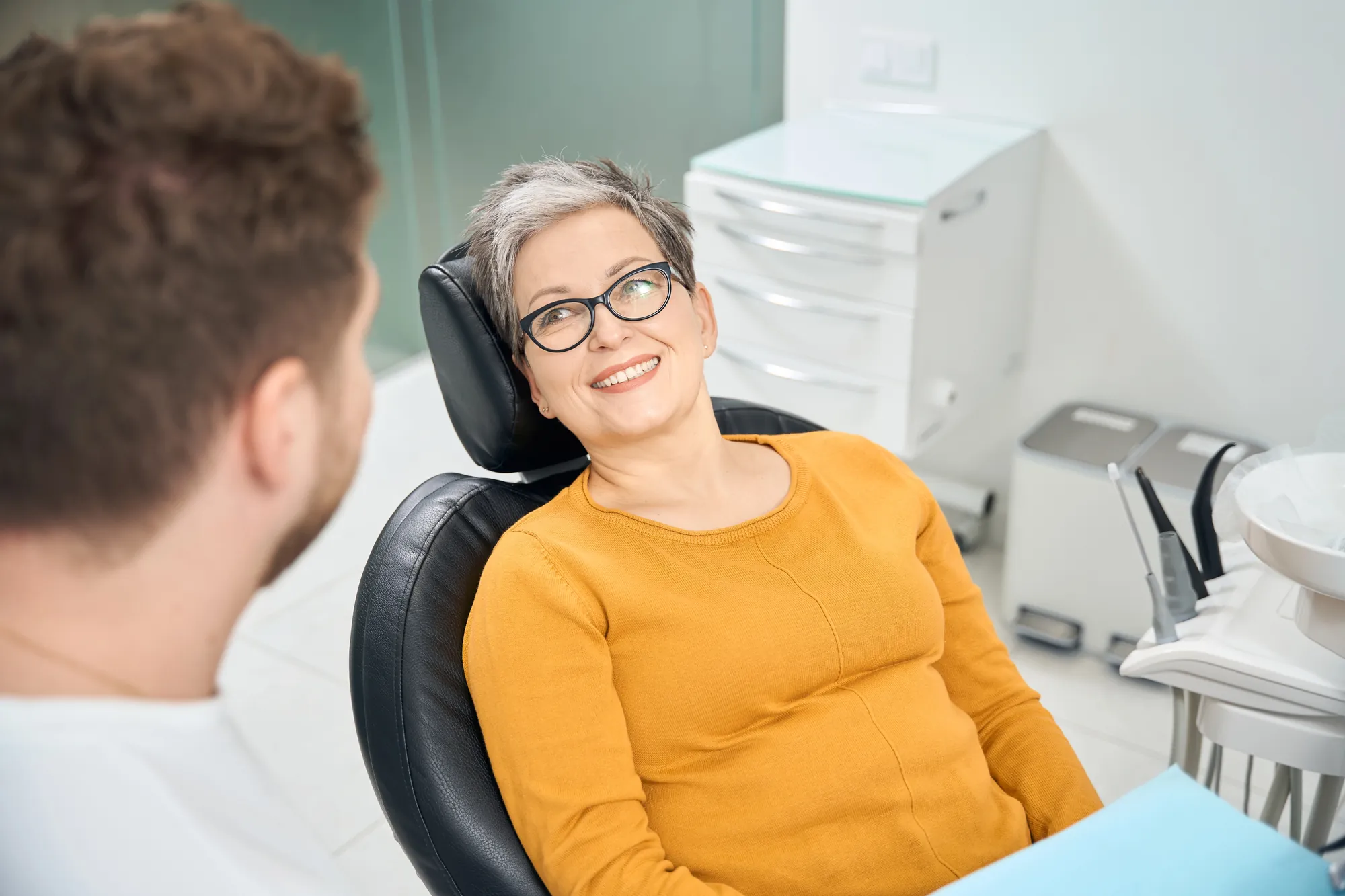 Smiling middle-aged woman with glasses sitting in a dental chair talking to a dentist.