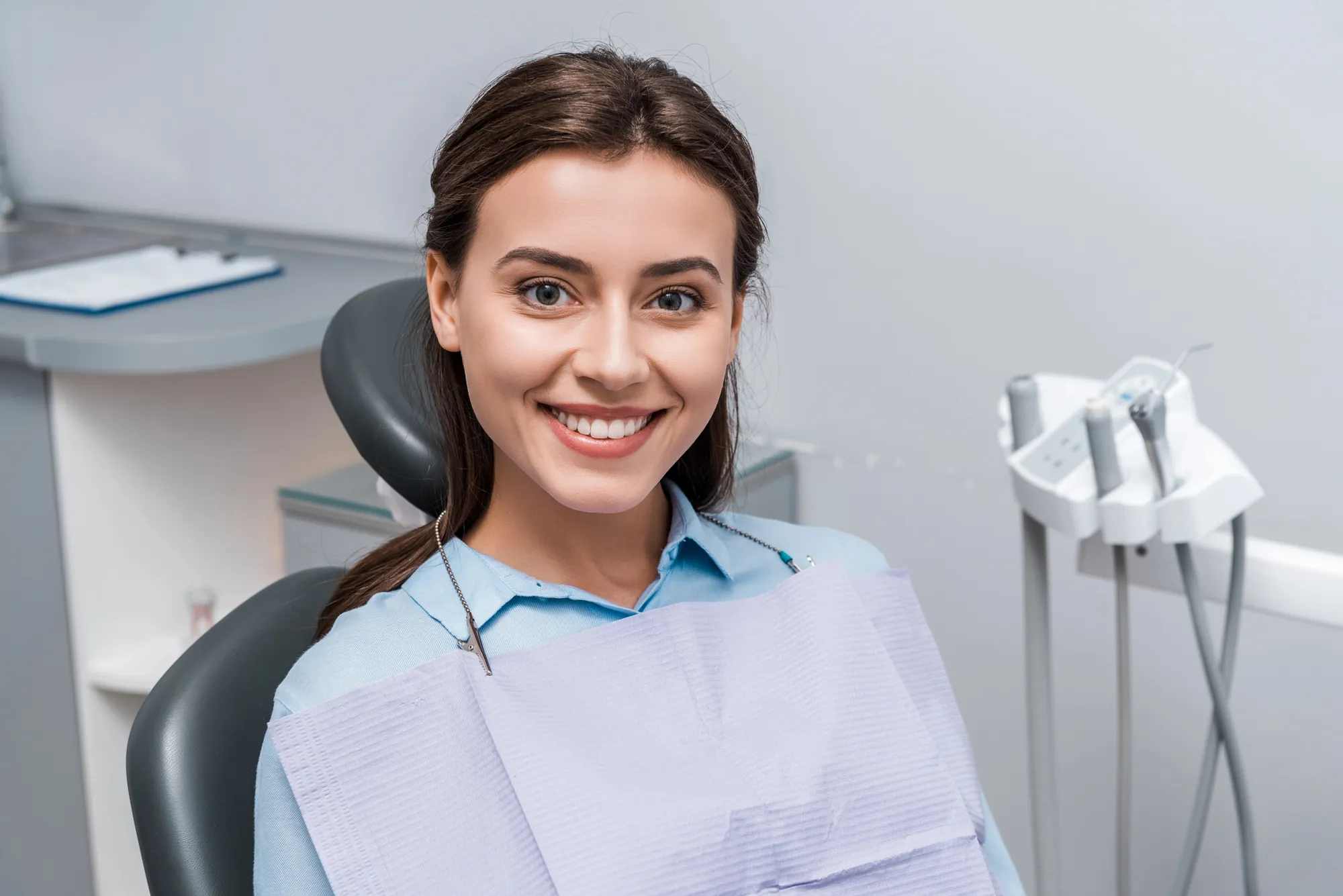 Smiling young woman sitting in a dental chair wearing a light blue shirt and a dental bib.