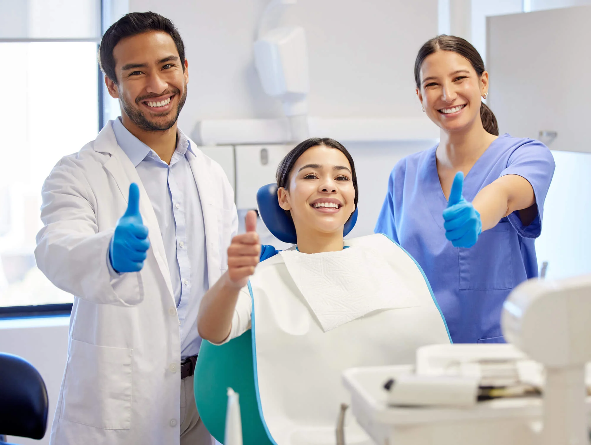Smiling dentist, patient, and dental assistant all giving thumbs up in a bright dental clinic.