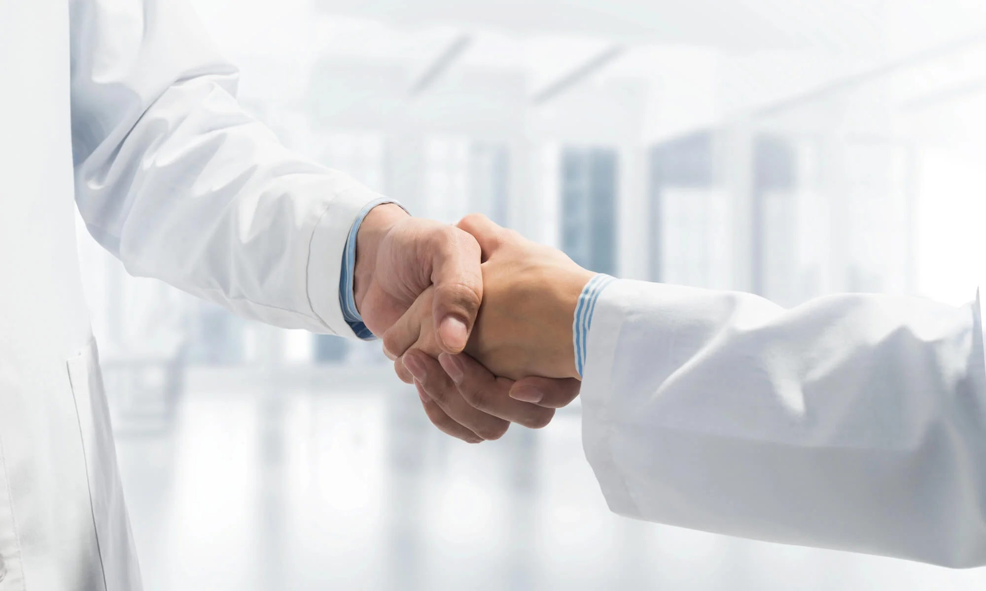 Two medical professionals in white coats shaking hands in a bright clinical setting.