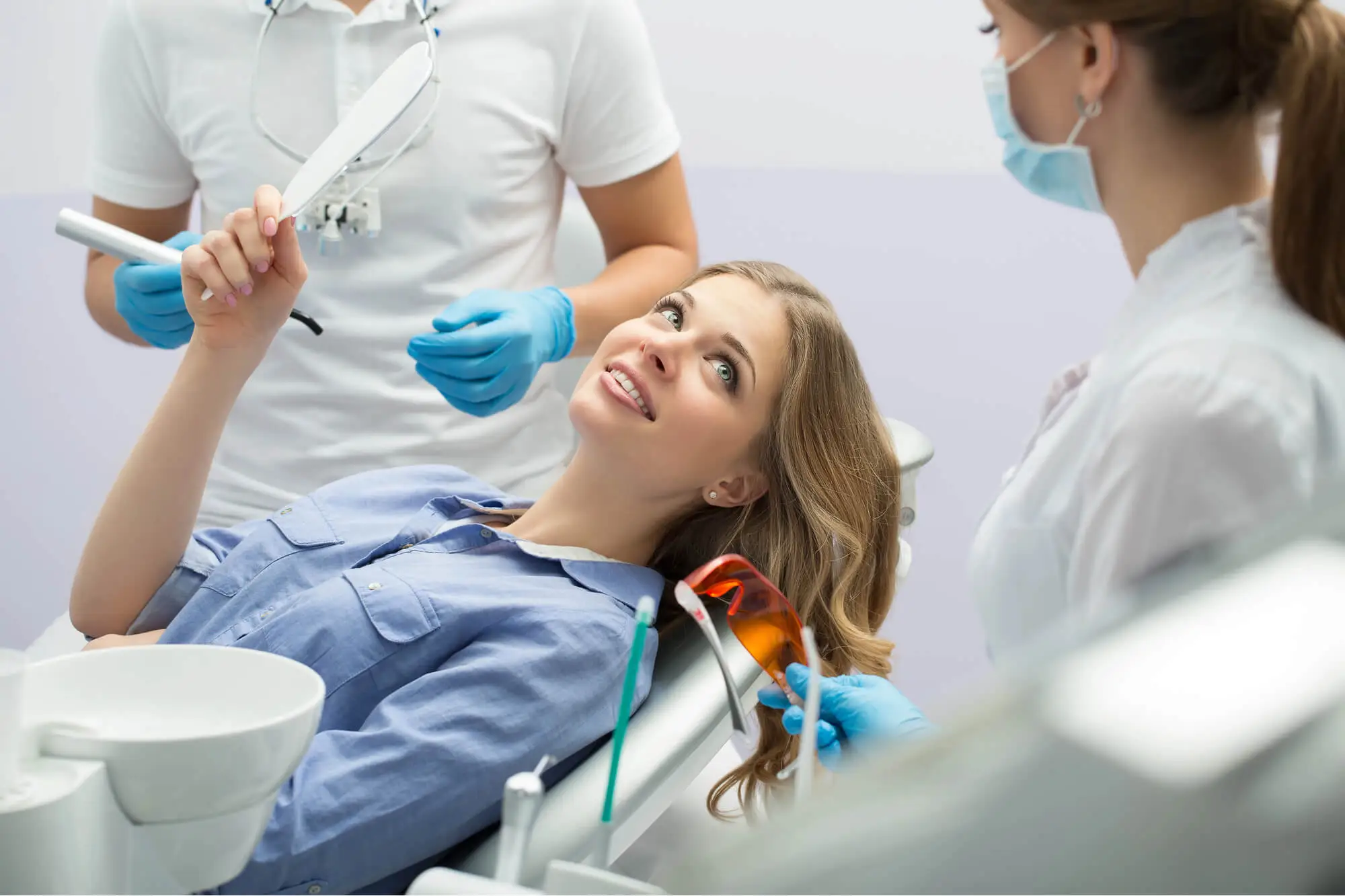 Woman in dental chair holding a mirror, looking at two dental professionals wearing masks and gloves.