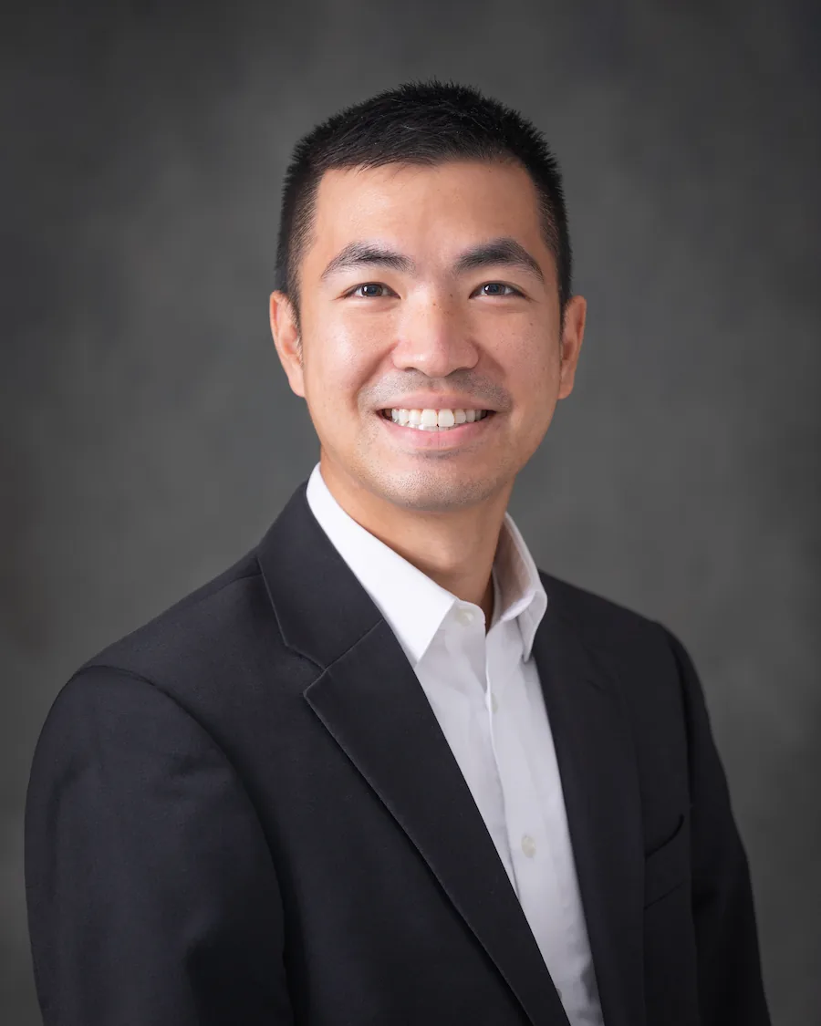 Smiling man with short black hair wearing a black suit jacket and white dress shirt against a gray background.