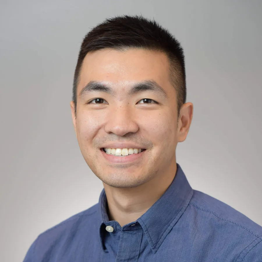 Smiling Asian man wearing a blue button-up shirt in front of a plain light gray background.