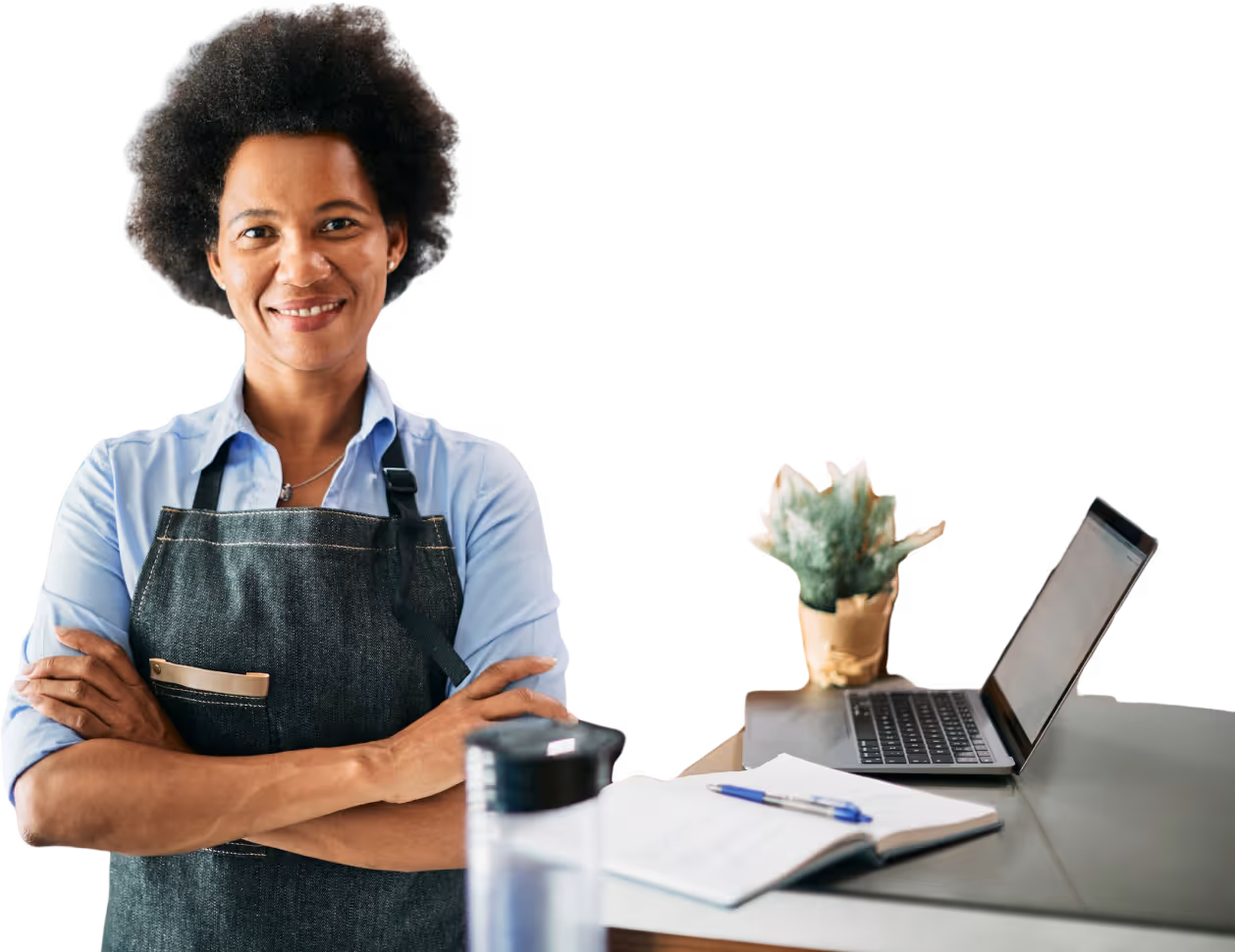 Une femme avec le sourire à côté d'un bureau sur lequel est posé un macbook et un cahier de note.
