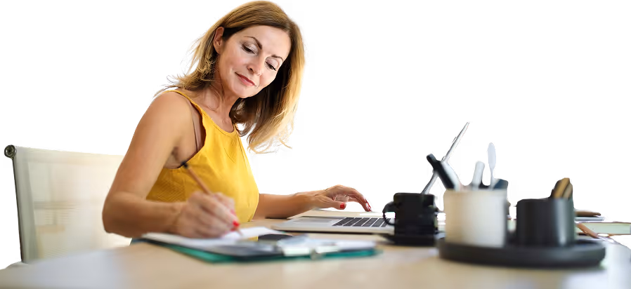 Une femme en train de travailler. Un ordinateur à sa gauche et des carnets à sa droite avec un stylo dans sa main droite.