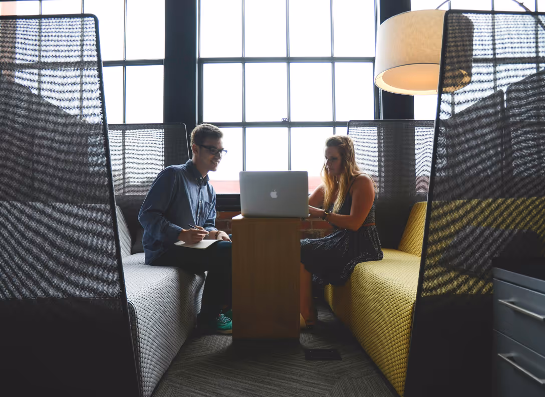 Deux jeunes adultes collaborent dans un espace de travail moderne, assis sur des canapés, un ordinateur portable posé sur une petite table. De grandes fenêtres laissent entrer la lumière naturelle.