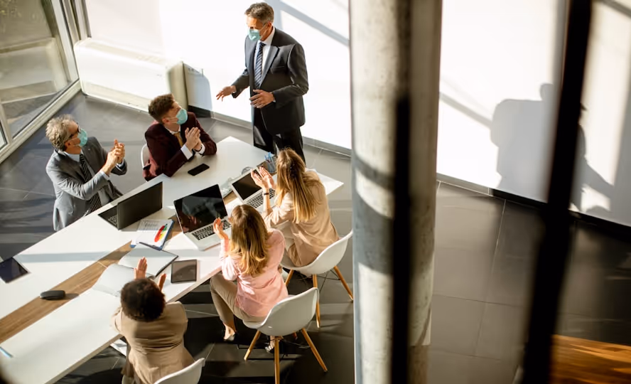 Un groupe de professionnels portant des masques participe à une réunion autour d'une table avec des ordinateurs portables, dans un bureau moderne bien éclairé.