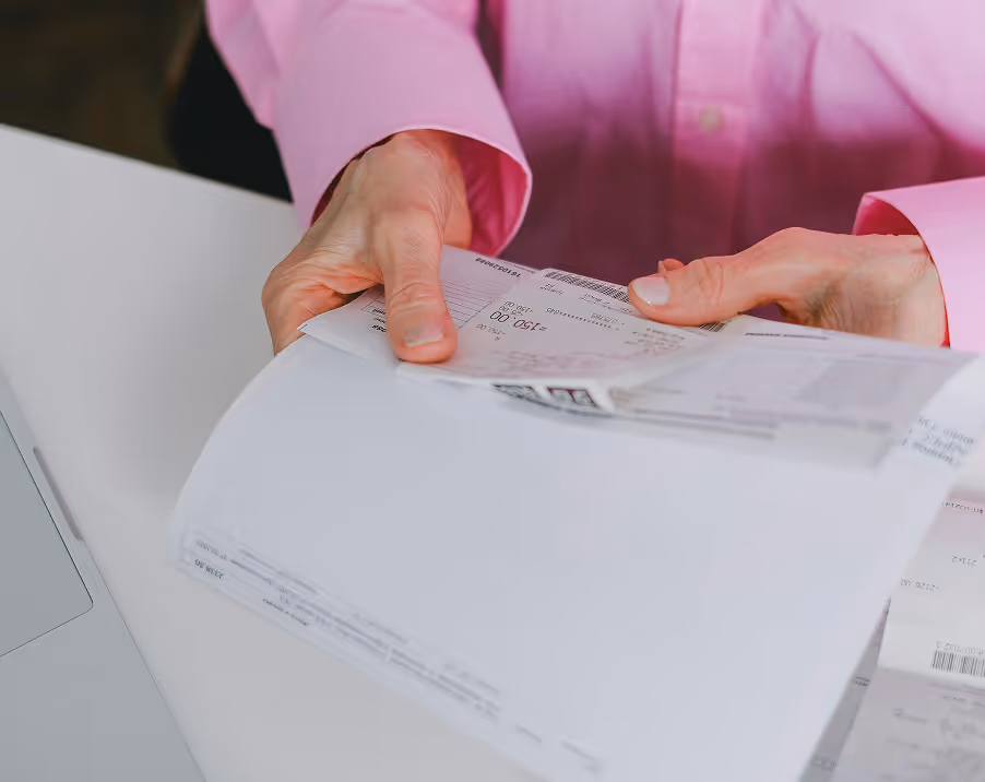 Une personne vêtue d'une chemise rose tient des reçus et des documents, avec un ordinateur portable partiellement visible à l'arrière-plan.