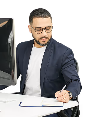 Homme en blazer prenant des notes à un bureau équipé d'un ordinateur, concentré et absorbé par son travail.