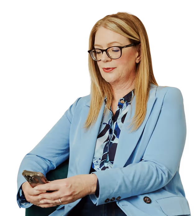 Une femme vêtue d'un blazer bleu clair est assise et regarde son smartphone. Elle a les cheveux longs et raides et porte des lunettes, ce qui lui donne une allure professionnelle.