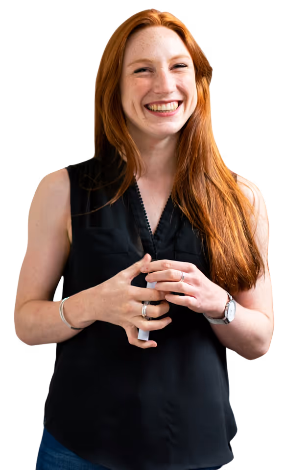 Une femme souriante aux longs cheveux roux se tient debout, tenant un petit objet, vêtue d'un haut noir sans manches et de bijoux en argent.