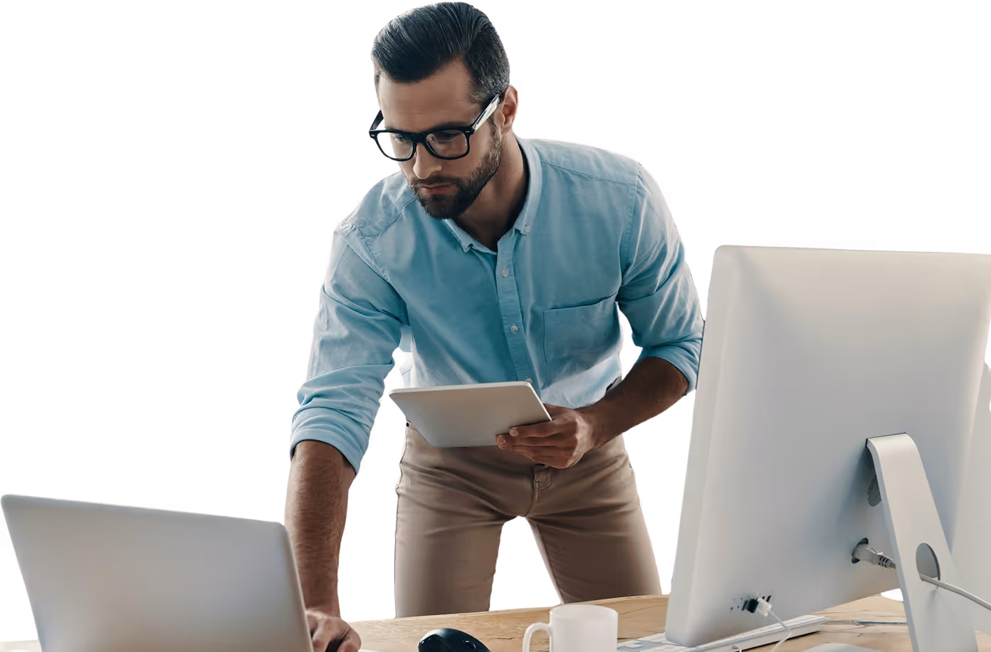 Un homme vêtu d'une chemise bleu clair se tient debout devant un bureau, tenant une tablette, travaillant sur un ordinateur portable et un ordinateur de bureau.