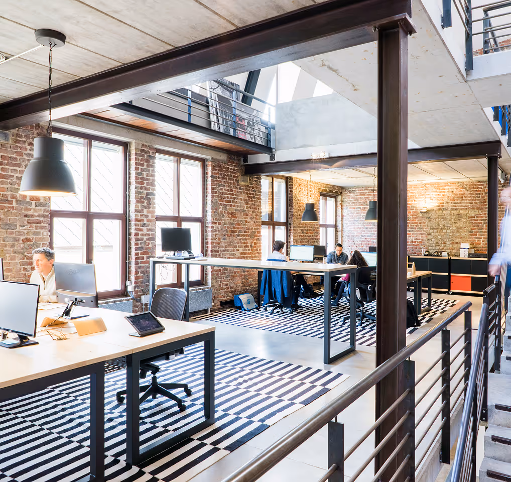 Espace de bureau moderne avec murs en briques, grandes fenêtres et postes de travail ouverts équipés d'ordinateurs et d'un tapis rayé.