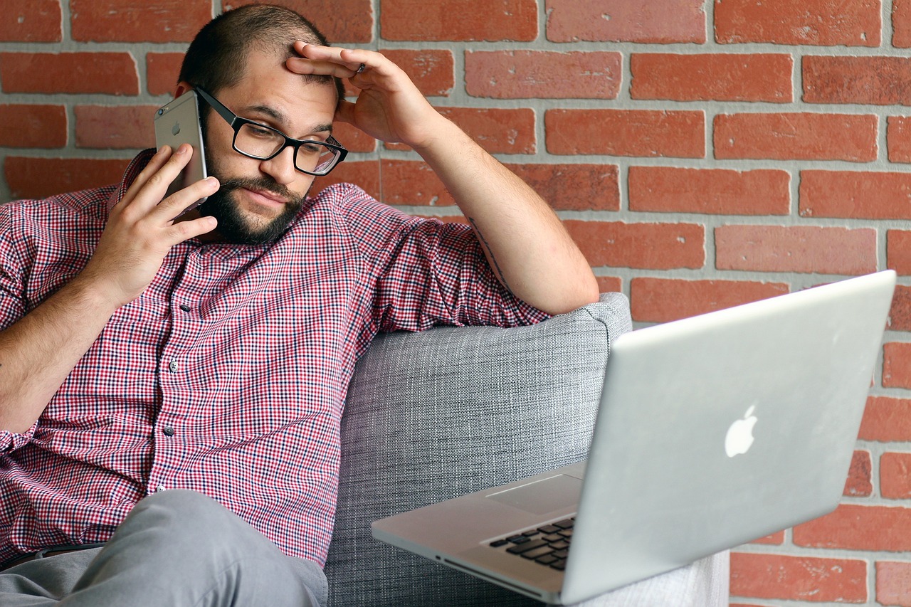 A concerned man in glasses, on a phone call, looking at his laptop with a hint of stress, possibly discussing contractor termination details, with a backdrop of a brick wall adding to the serious atmosphere of the conversation.