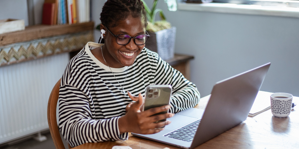 Cheerful woman wearing glasses, earphones, and a striped shirt multitasking with a smartphone and laptop in a home office, embodying the dynamic lifestyle of remote company professionals.