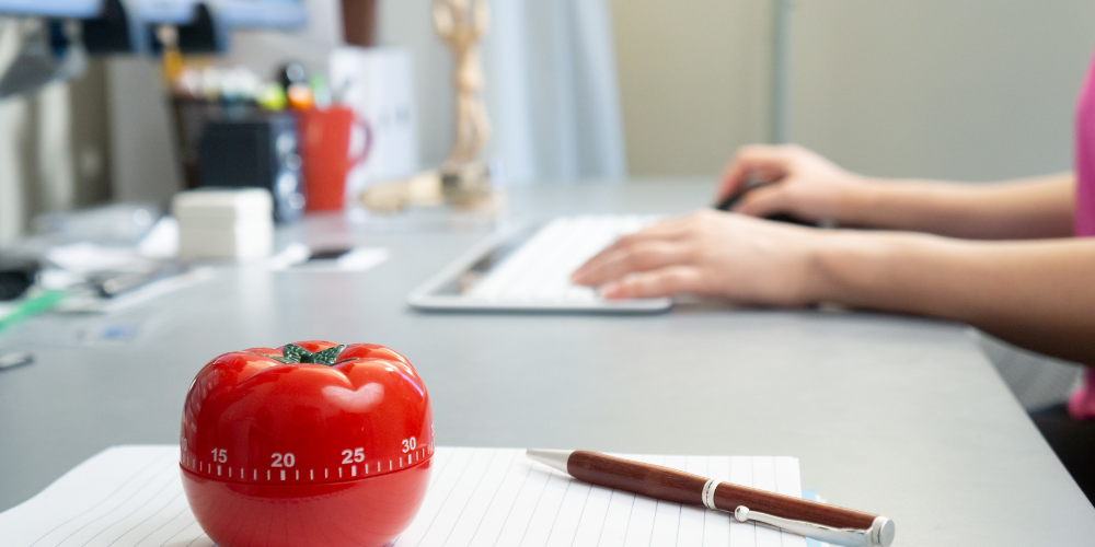 Focused work environment with a tomato-shaped Pomodoro timer on a notepad, alongside a professional remotely working on a computer, embodying time management practices in a remote company.