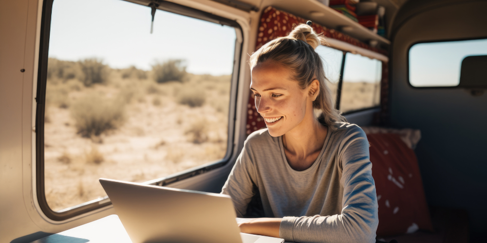 A woman in a sunlit van smiles while working on her laptop, exemplifying the lifestyle of a digital nomad surrounded by a desert landscape, embracing the freedom of remote work