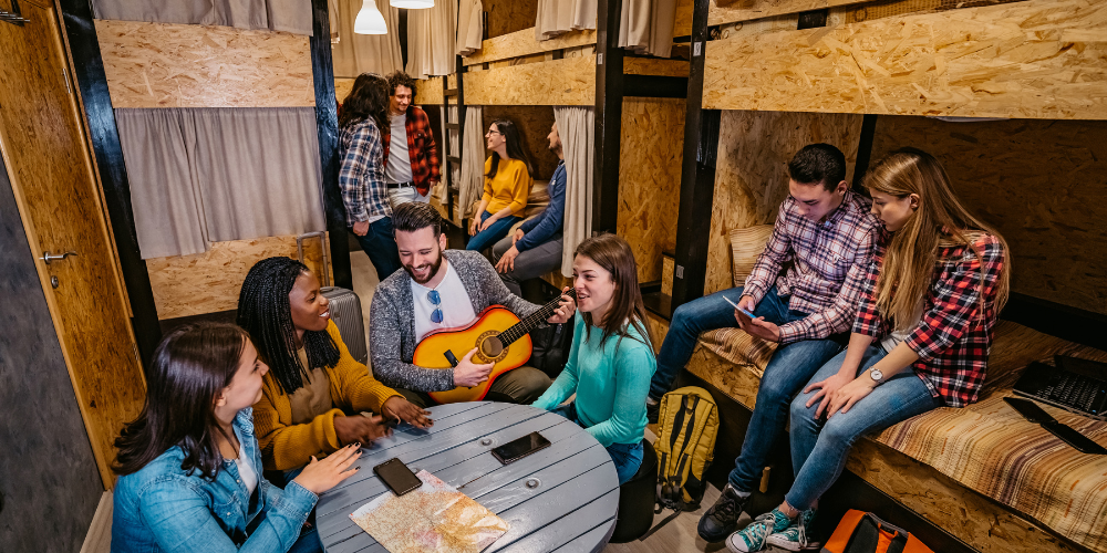Group of young, joyful people in a cozy hostel common area, with one playing the guitar, embodying the community and shared experiences of digital nomad life.