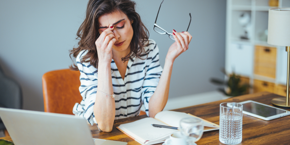 A stressed professional woman massaging her temples, reflecting the challenges of termination of employment, with her work notebook, glasses in hand, and laptop on the table.