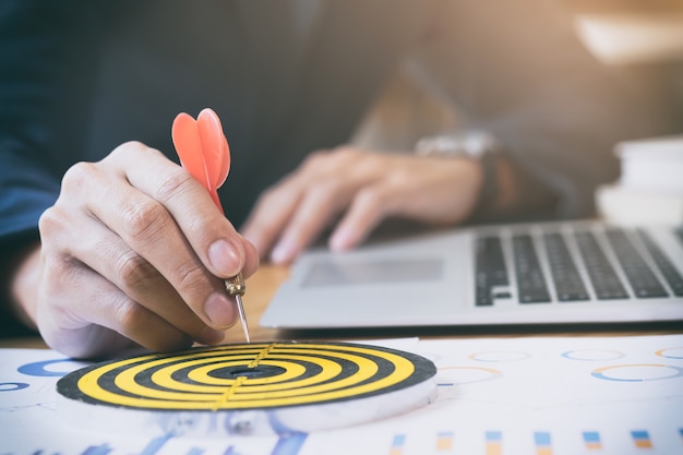 The image displays a person's hand placing a red dart onto the center of a target bullseye, which is on top of a desk with charts, graphs, and a laptop in the background. This conveys a concept of goal achievement, precision, or reaching marketing targets, which is relevant to the performance evaluation of an offshore social media manager.