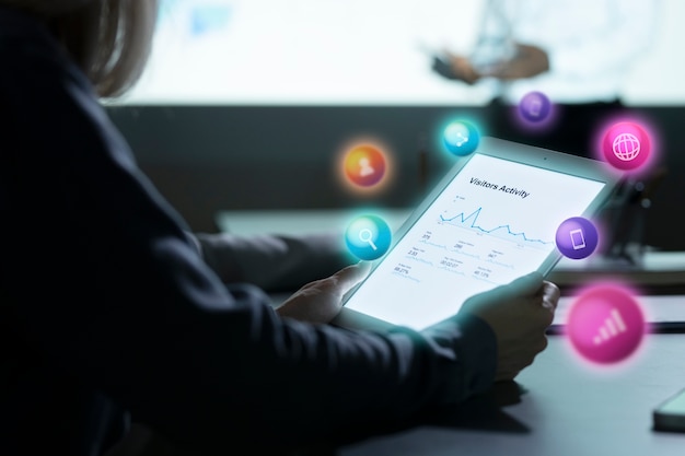 The image features a person holding a tablet displaying a graph labeled "Visitors Activity", with various analytics or performance metrics visible on the screen. Colorful icons suggestive of social media and web functions are floating around the tablet, indicating digital analytics or social media management activities, which would be relevant to the theme of monitoring and evaluating the performance of an offshore social media manager.