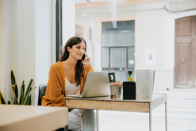 A woman sits at a desk in a bright office, speaking on a mobile phone with a laptop open in front of her, accompanied by a water bottle and a small potted plant, representing an SEO Specialist at work.