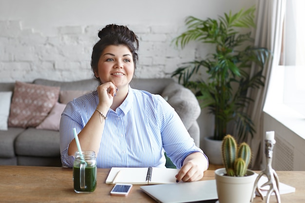 A cheerful woman with a bun hairstyle sitting at a wooden desk in a casual workspace with a striped blue shirt, taking notes next to a laptop, a green smoothie, and a cactus plant, suggesting a healthy and organized work environment.