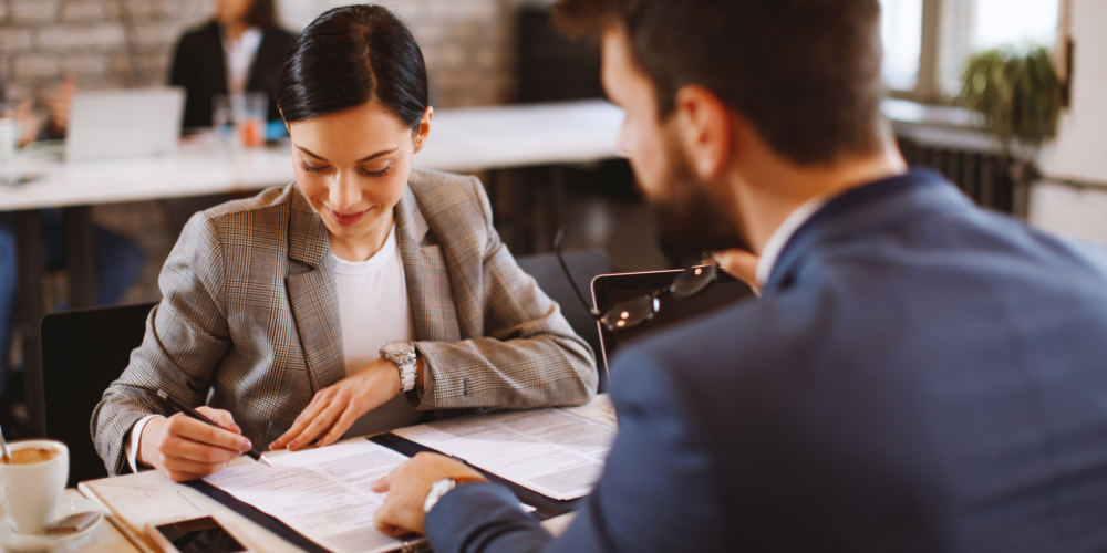 A businesswoman attentively signs a document across from a male colleague in a bustling office environment, capturing a moment of professional agreement or contract finalization.