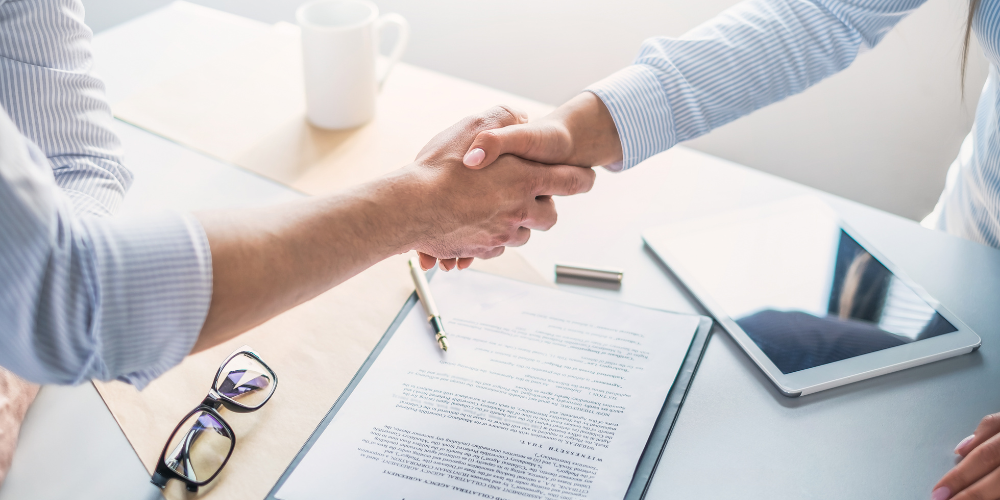 Two individuals engaging in a firm handshake over a desk with a signed document, glasses, and a tablet, conveying a professional agreement or conclusion of a business meeting.