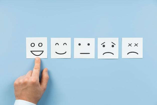 A hand pointing to the happiest face in a line-up of four square cards, each with a different facial expression ranging from happy to sad, against a blue background.