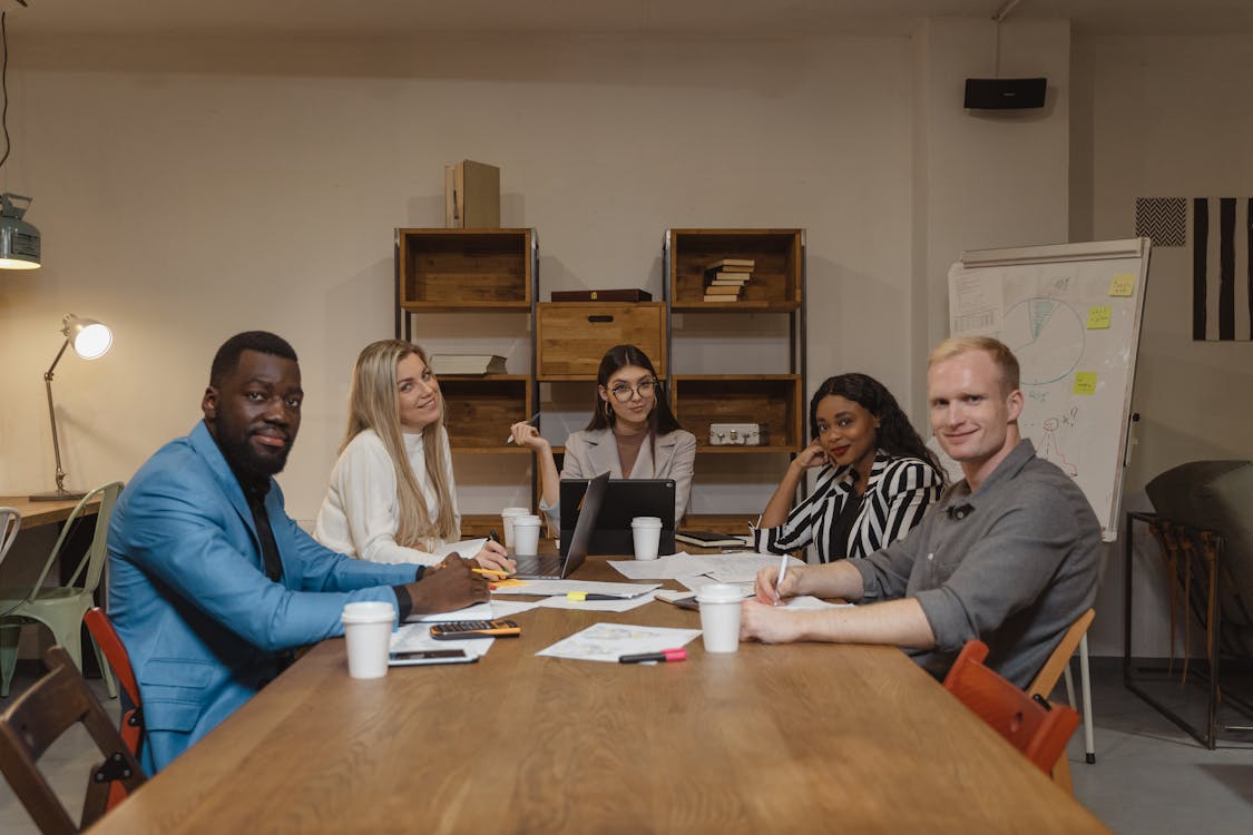 A diverse team of five seated around a meeting table, with papers and laptops, smiling and engaging in a collaborative discussion in a modern office setting.