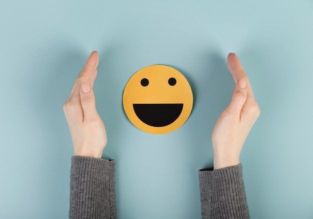 Two hands framing a smiling emoji face against a pastel blue background.