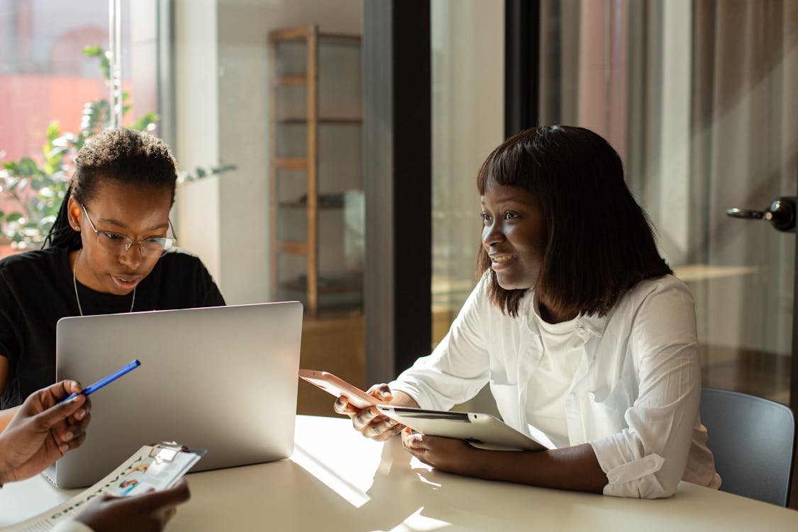 Two professionals engaged in a discussion with one looking at a laptop and the other holding a tablet, in a brightly lit office space.