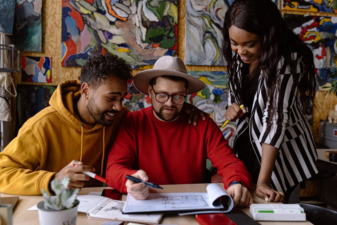 Three diverse team members collaborating over a notebook with one pointing at a document, in a creative workspace adorned with colorful paintings.