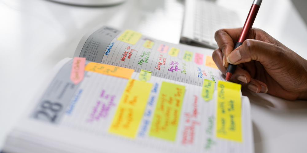 A person's hand writing on a planner with colorful sticky notes and highlighter marks, organizing tasks and appointments, showcasing meticulous schedule management.