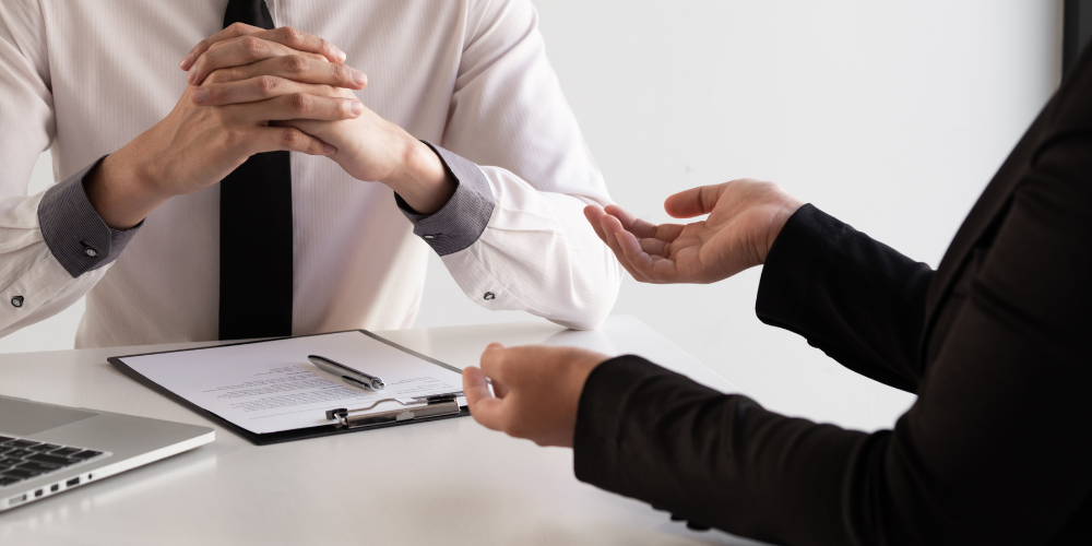 Two individuals engaged in a formal discussion, one gesturing explanatorily, with a document and a laptop on the table, suggesting a business negotiation or interview setting.
