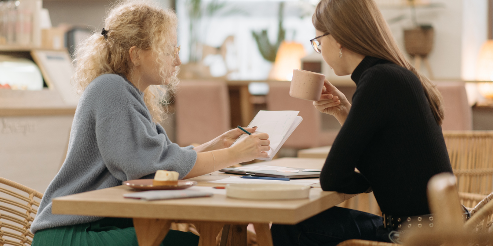 Two women engaged in a focused discussion at a cafe, with one holding papers and the other sipping a drink, amidst a relaxed yet productive atmosphere.