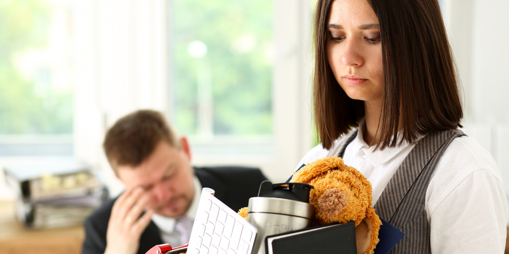 A young woman carrying office items, including a teddy bear and a coffee mug, looks downhearted, possibly reflecting job loss or the emotional moment of leaving a workplace.