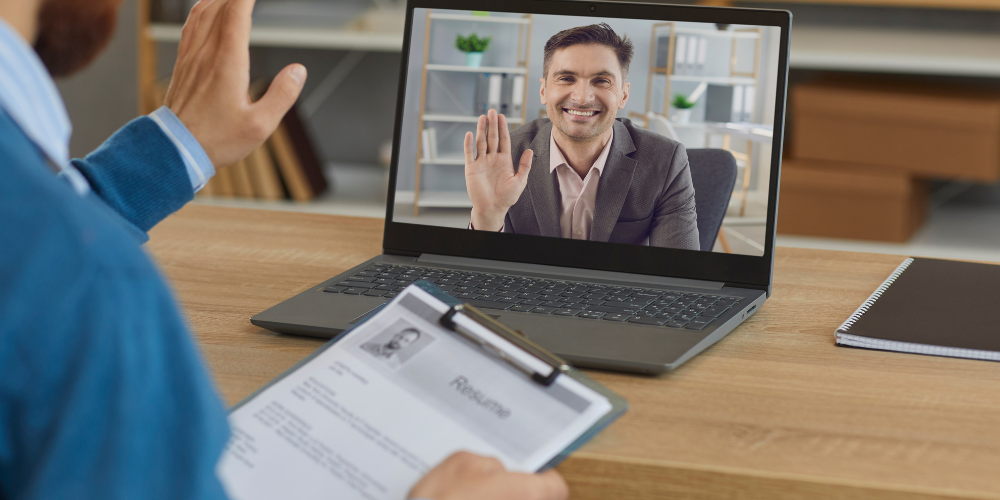 A person in a blue sweater is participating in a video call with a smiling man on the screen, who is waving hello. There's a resume on the table in front of the person, indicating a possible job interview or meeting scenario.