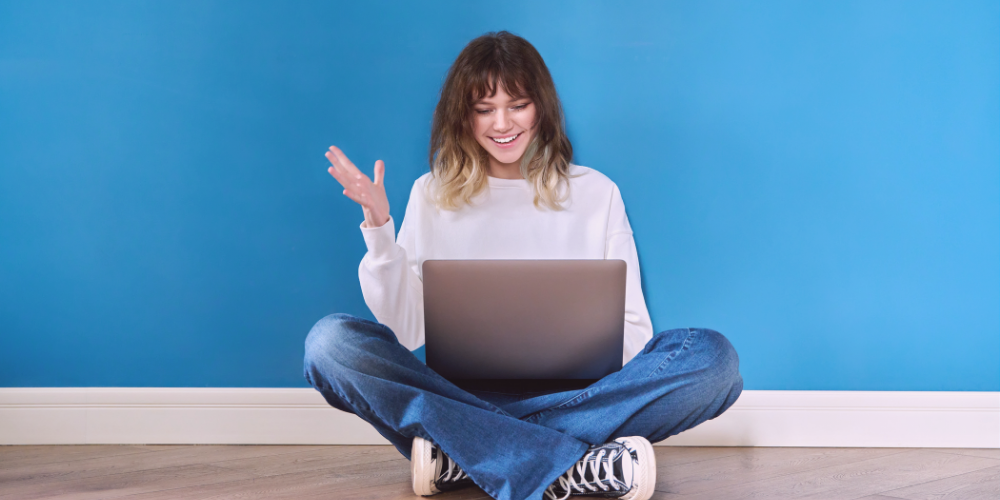 A woman in casual attire sits cross-legged on the floor with a laptop, smiling and gesturing with her hand, likely engaged in a friendly virtual conversation. The room has a blue wall and wooden floor.