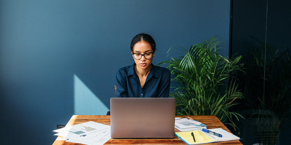 A focused woman in glasses working at a desk with a laptop, papers, and a notebook in a room with a dark wall and potted plants, reflecting a serious and professional remote work environment.