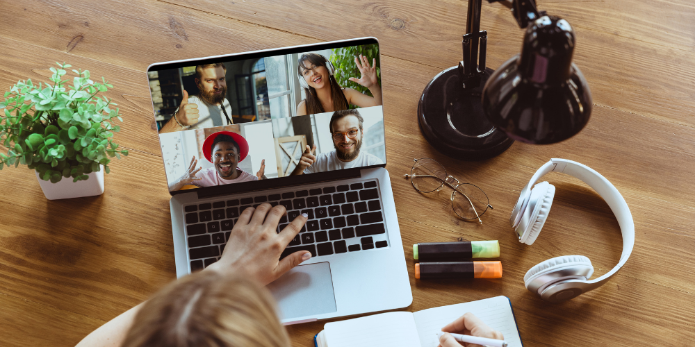 An overhead view of a person's hands typing on a laptop with a video call on the screen, showing four happy participants. The desk is also equipped with a desk lamp, headphones, a notepad, a potted plant, and glasses, representing a well-organized remote work setup.