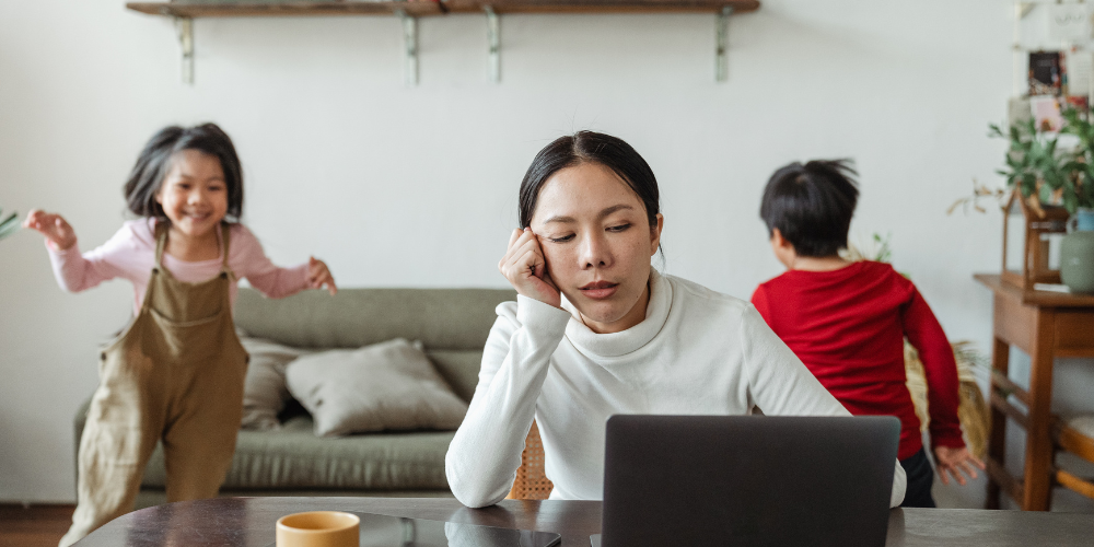 A woman in a white sweater looks tired or stressed while working on a laptop at a wooden table, hand to forehead. In the background, two children play, one in a pink outfit mid-dance move and another in red, suggesting the multitasking nature of remote work and family life.