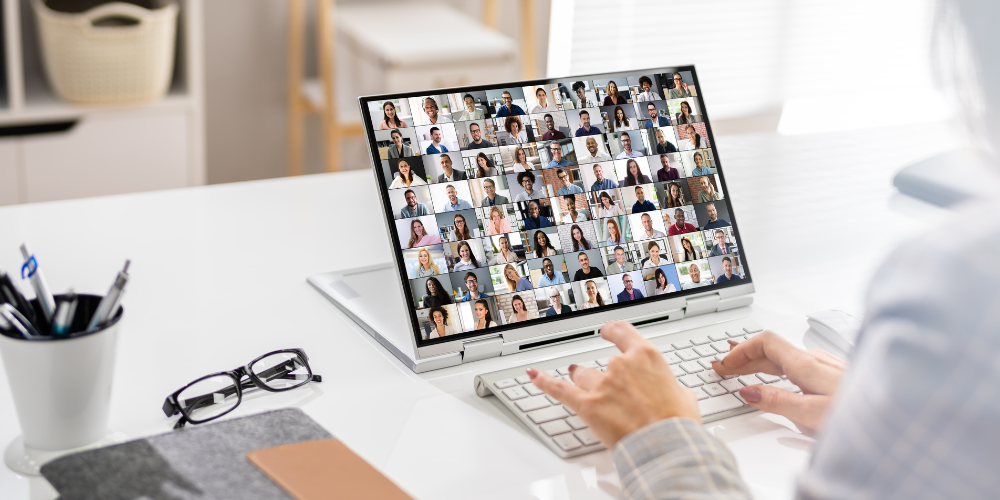 Rear view of a person working on a laptop displaying a large virtual meeting screen with many participants, depicting a common scene in remote and hybrid work environments.