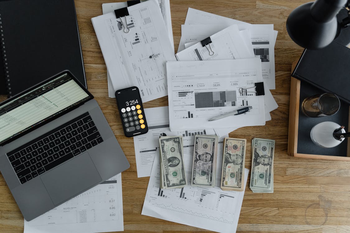 Top view of a workspace with a laptop, calculator on smartphone, financial documents, and US dollar bills, depicting finance management.